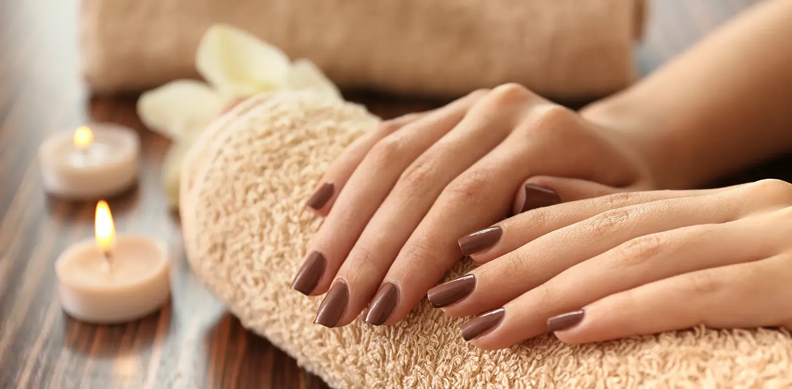 Short nails in glossy chocolate brown, hands resting on a rolled towel beside a candle